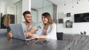 Pareja sonriendo felices sentados frente a la pantalla de su laptop en su apartamento rentado