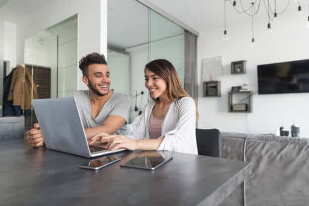 Pareja sonriendo felices sentados frente a la pantalla de su laptop en su apartamento rentado