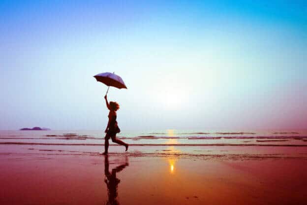 Mujer corriendo feliz junto al mar sosteniendo una sombrilla de color violeta frente al atardecer