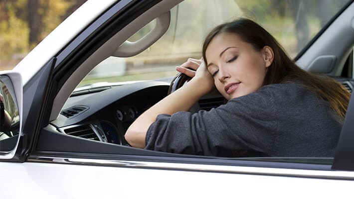 Mujer joven recostada con los ojos cerrados sobre el volante de su auto conduciendo estando cansada