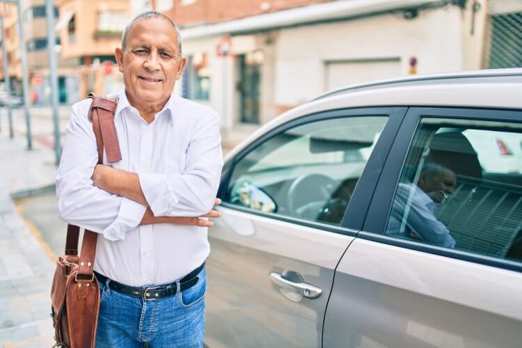 hombre latino mayor sonriendo frente a su auto con seguro basico
