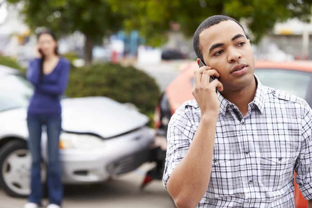 hombre hablando por celular en primer plano con un accidente de auto de fondo