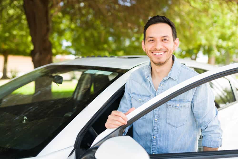hombre hispano sonriendo y posando con su auto con seguro barato