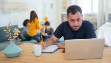 man looking at laptop and taking notes with this kids in the background