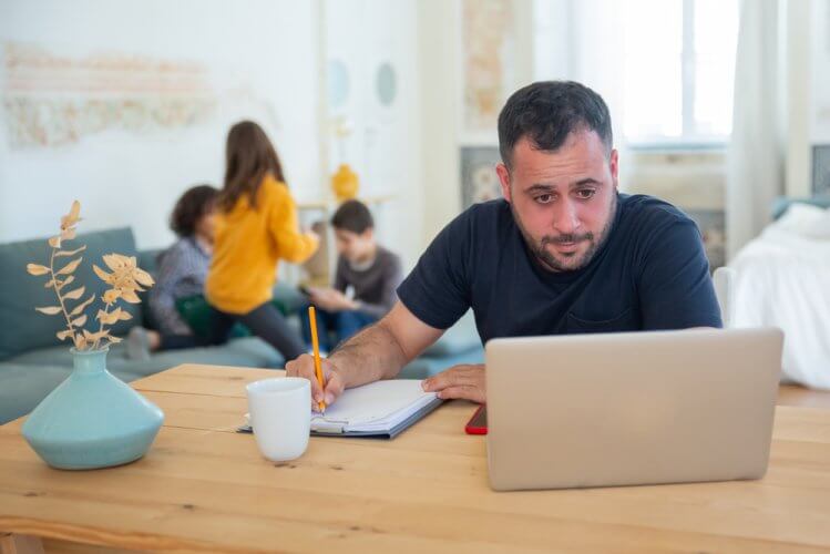 man looking at laptop and taking notes with this kids in the background