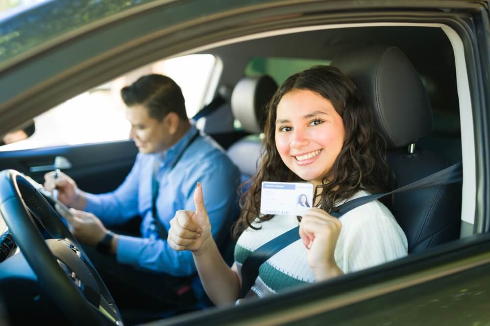 Estudiante sonriente muestra su licencia y hace una señal de aprobación dentro del auto después de practicar cómo aprobar la prueba de manejo en USA.