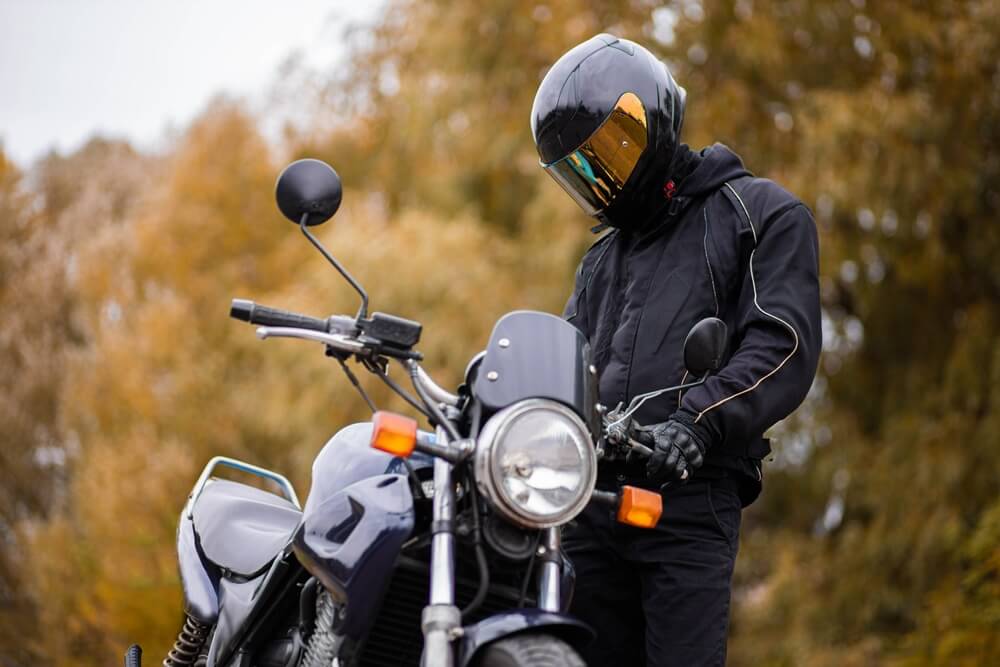 Motociclista con casco revisa su moto detenida en un camino rodeado de árboles, ideal para aprender cómo manejar una motocicleta de cambios.