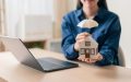 Una mujer sostiene una casa en miniatura protegida con un paraguas junto a una laptop, ilustrando la comparación entre cobertura amplia vs limitada en seguros