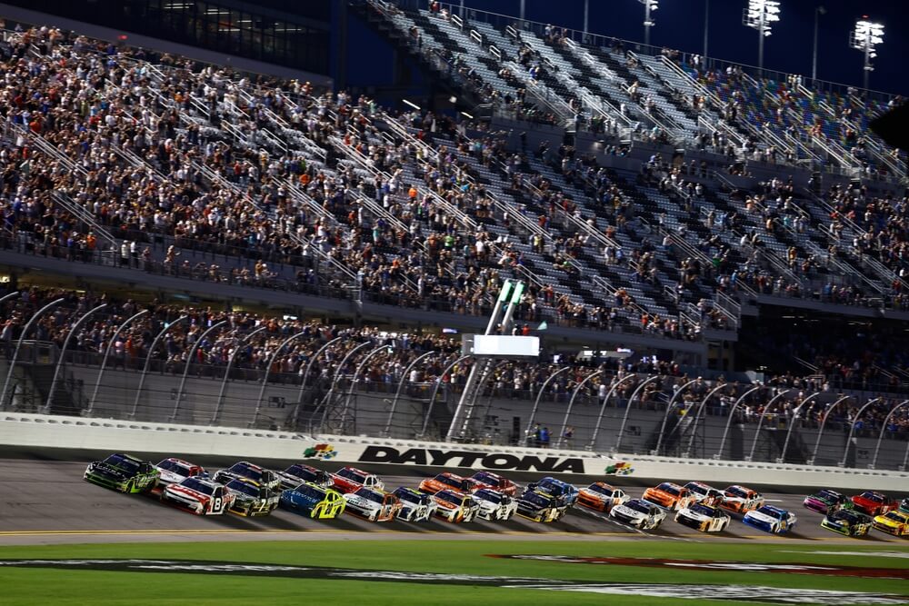 Autos compiten lado a lado en un óvalo iluminado con gradas llenas, mostrando la intensidad de las carreras de NASCAR de noche.