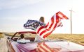 Pareja viajando en un auto convertible por carretera en Estados Unidos mientras sostienen una bandera estadounidense al viento.