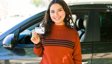 Mujer joven sonriente sosteniendo su licencia de conducir frente a su automóvil, celebrando haber obtenido su licencia en Estados Unidos.