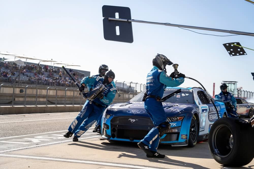 Equipo en pits cambia llantas y recarga combustible durante carrera, mostrando cómo se prepara un piloto de NASCAR