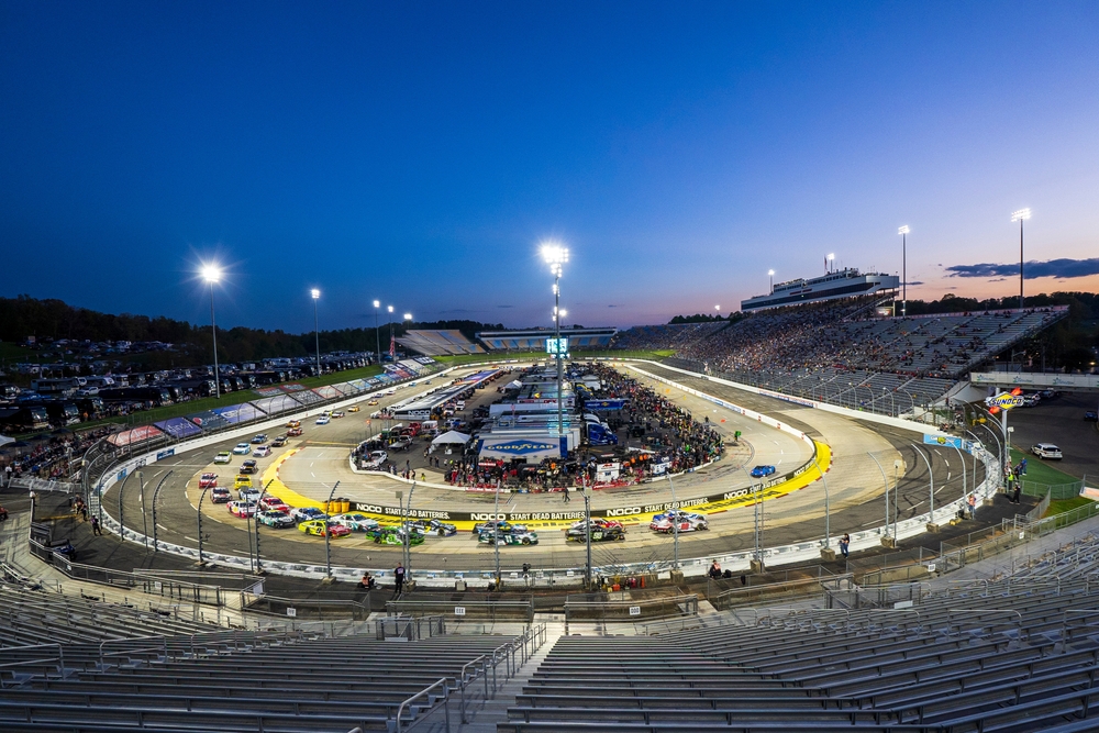 Vista aérea de circuito oval iluminado con autos compitiendo y público, representando las pistas icónicas de NASCAR.