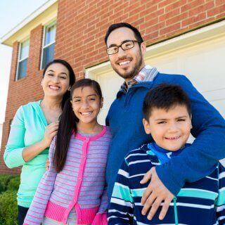 Familia de cuatro personas abrazados y sonriendo felices frente a su casa de ladrillos