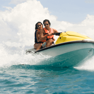 Hombre joven de gafas oscuras con chaleco conduciendo moto de agua en el mar junto a chica sonriente