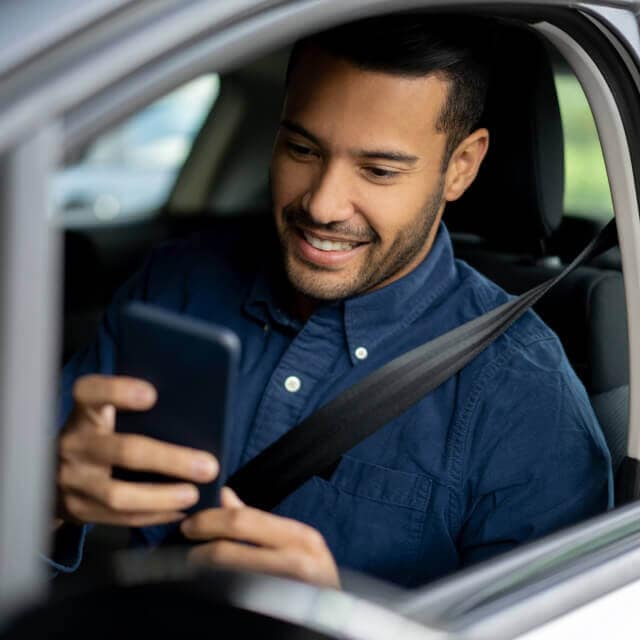 Hombre sonriente en el asiento de conductor de un auto viendo su celular