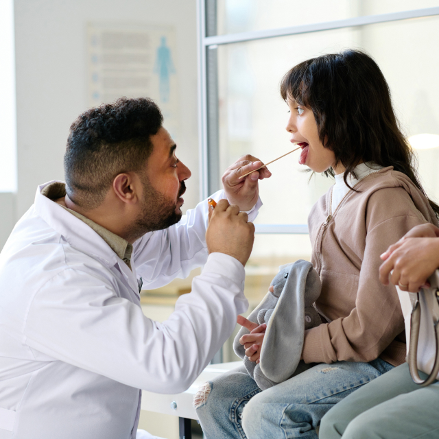 Médico latino juega con su estetoscopio con una niña paciente acompañada de su madre.