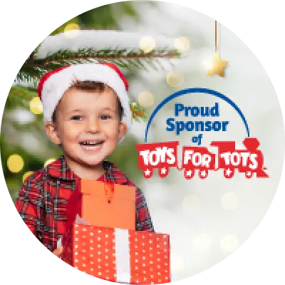 Un niño sonriente con camisa de cuadros rojos y gorro de Santa sostiene regalos envueltos frente a un árbol de Navidad nevado, junto al texto que dice “Orgulloso Patrocinador de Toys for Tots.”