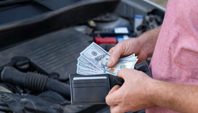 Persona sostiene dinero frente al motor de un automóvil, ilustrando el reto de cubrir reparaciones con ahorros propios en lugar de contar con un seguro de auto.